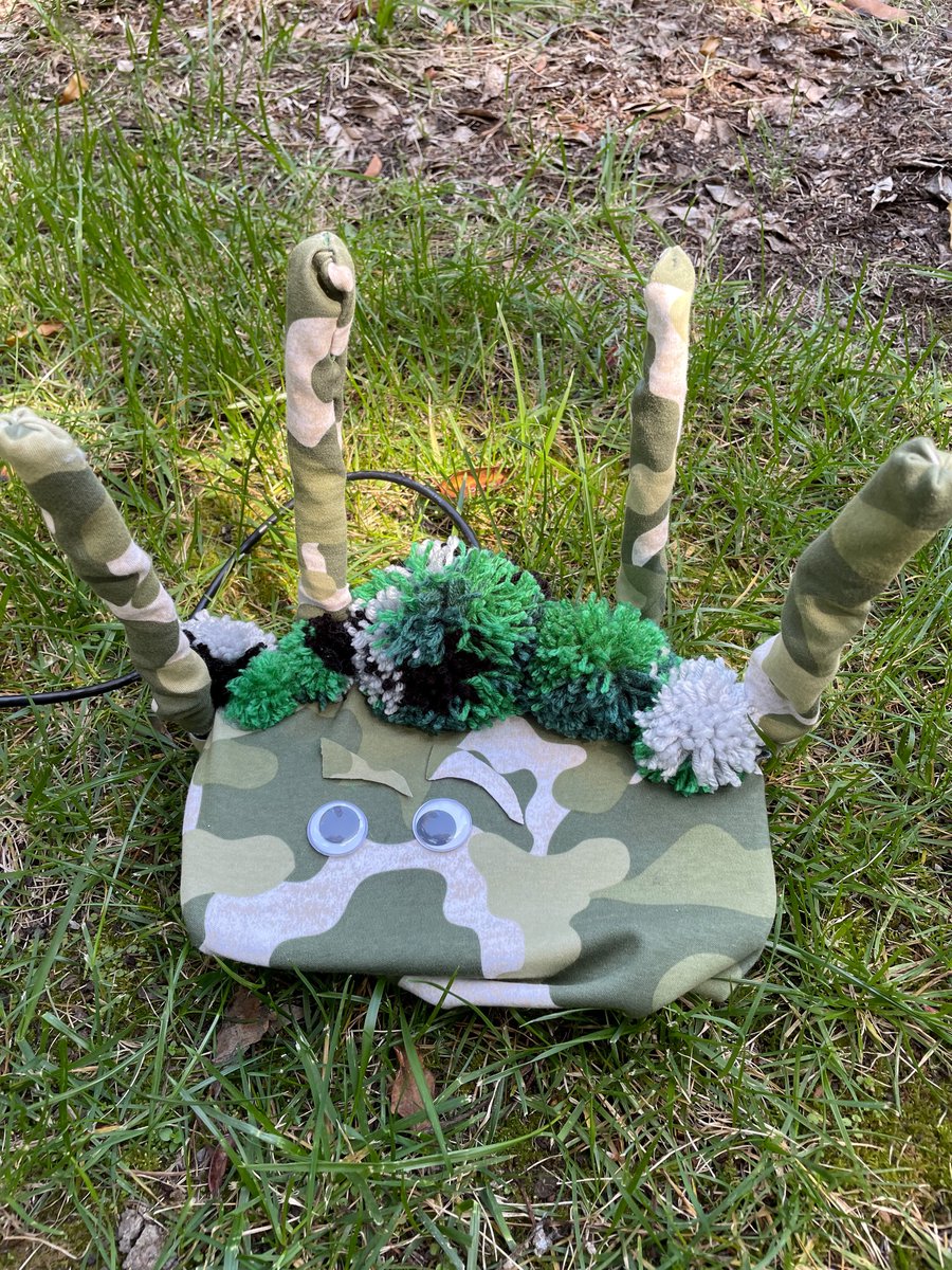 Picture of wifi router with camouflage, pom-poms, and googly eyes on top of grass with leaves in the background.