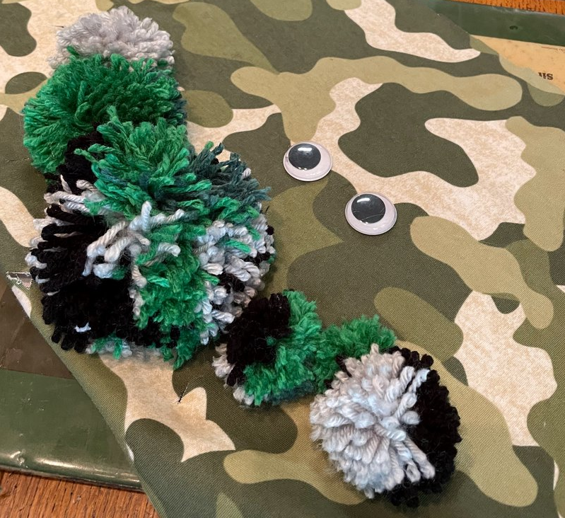 Picture of camouflage fabric with green/white/black yarn pom-poms and two googly eyes glued on.